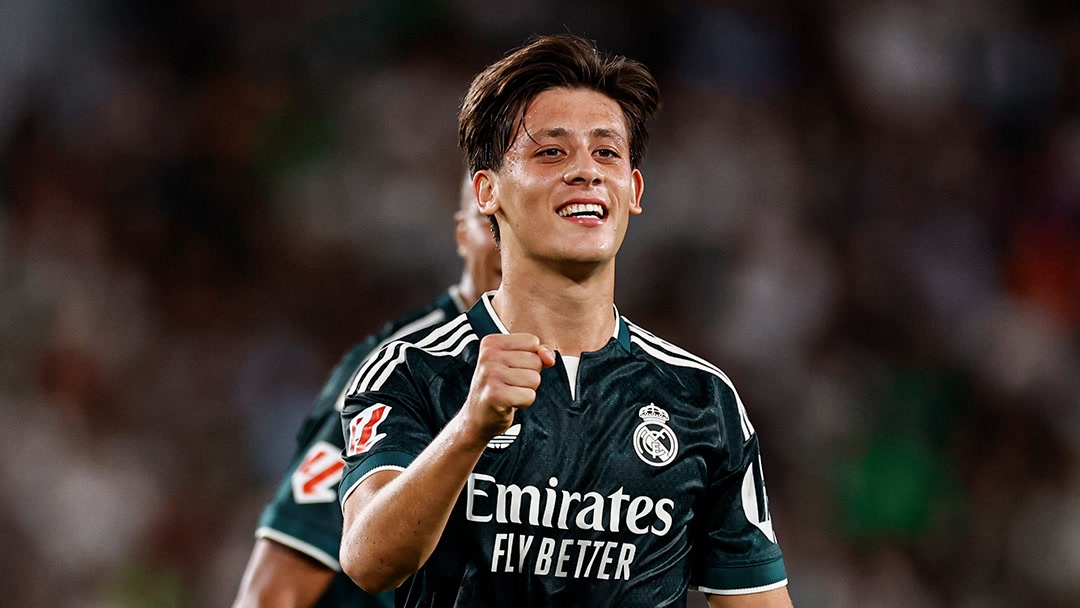 1080x608. Arda Güler Real Madrid deplasman formasıyla yakın plan, gülümseyip yumruk sıkıyor, arkada bulanık takım arkadaşı. Temiz, yazısız.
