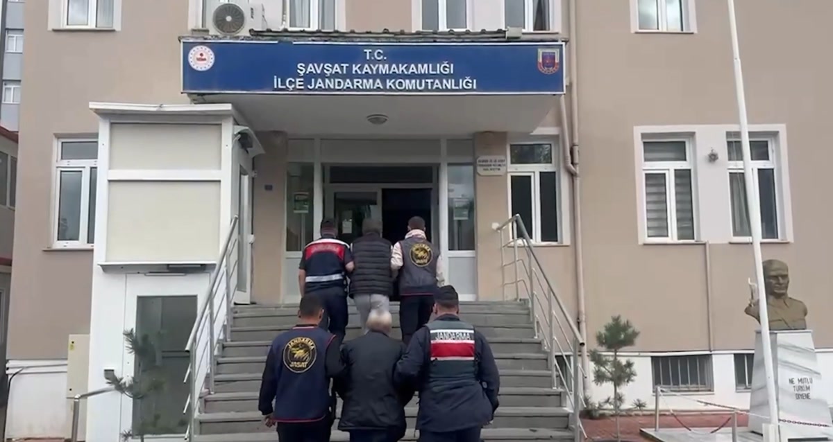 KAPAK ÖNERİSİ (1859x989): Şavşat Kaymakamlığı İlçe Jandarma Komutanlığı tabelalı binanın önü; JASAT yelekli jandarmalar zanlıyı merdivenlerden binaya götürüyor, yüzler arkadan. Temiz kare, yazı/kolaj yok.