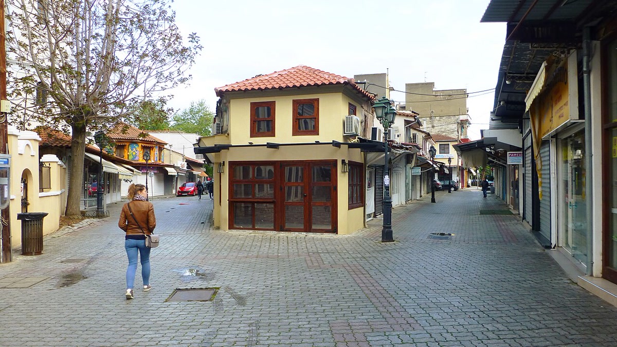 Gümülcine (Komotini) eski çarşısında kaldırım taşlı sokak, kiremit çatılı sarı bina ve esnaf dükkânları; yazısız, net sokak karesi (1280x721, CC BY-SA 4.0)