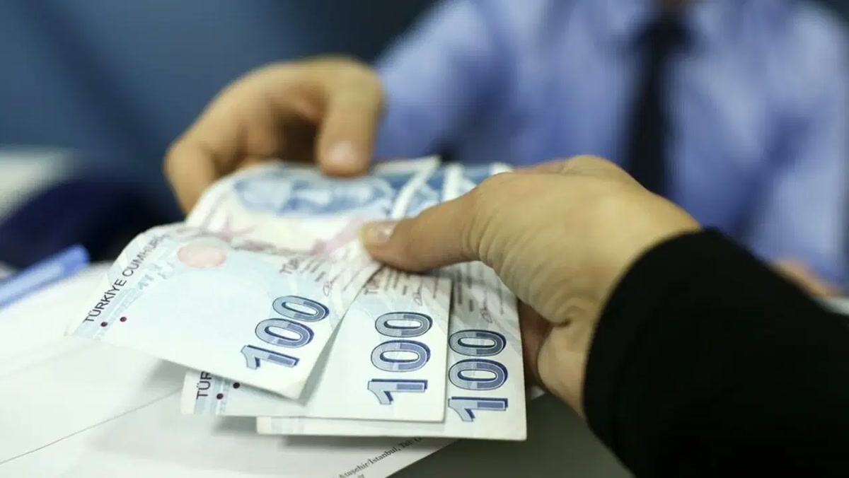 Gişe/ofis masasında bir elden diğerine uzatılan 100 TL banknotlar, arkada bulanık gömlekli yetkili (1280x720; sunucu .jpg adresine webp içerik döndürüyor).