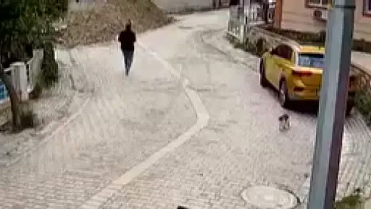 1280x720 güvenlik kamerası karesi: kadının kazadan hemen önce parke taşlı sokakta yürüdüğü an, sağda sarı taksi; logo yok, kamera kalitesinde hafif bulanık