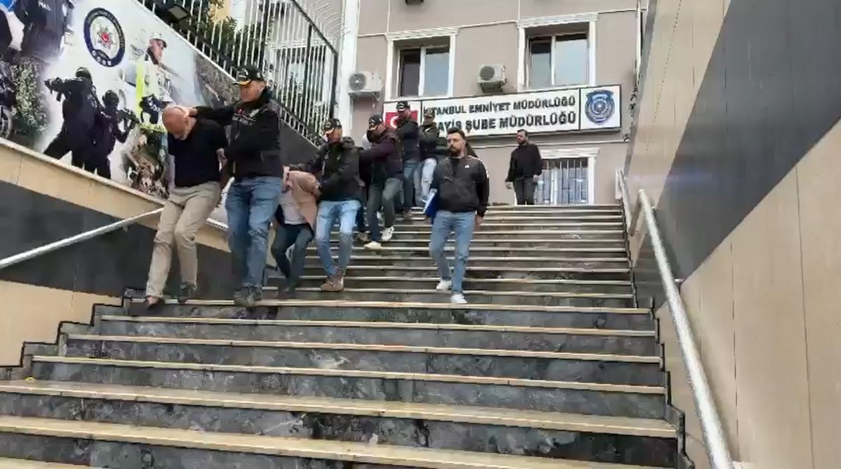 KAPAK ADAYI - Gözaltına alınan şüphelilerin İstanbul Emniyet Müdürlüğü binası önündeki merdivenlerden polis eşliğinde indirilişini gösteren gerçek operasyon fotoğrafı; yatay, net, olayın gerçek mekânı (emniyet binası) görünüyor, grafik/kolaj değil.