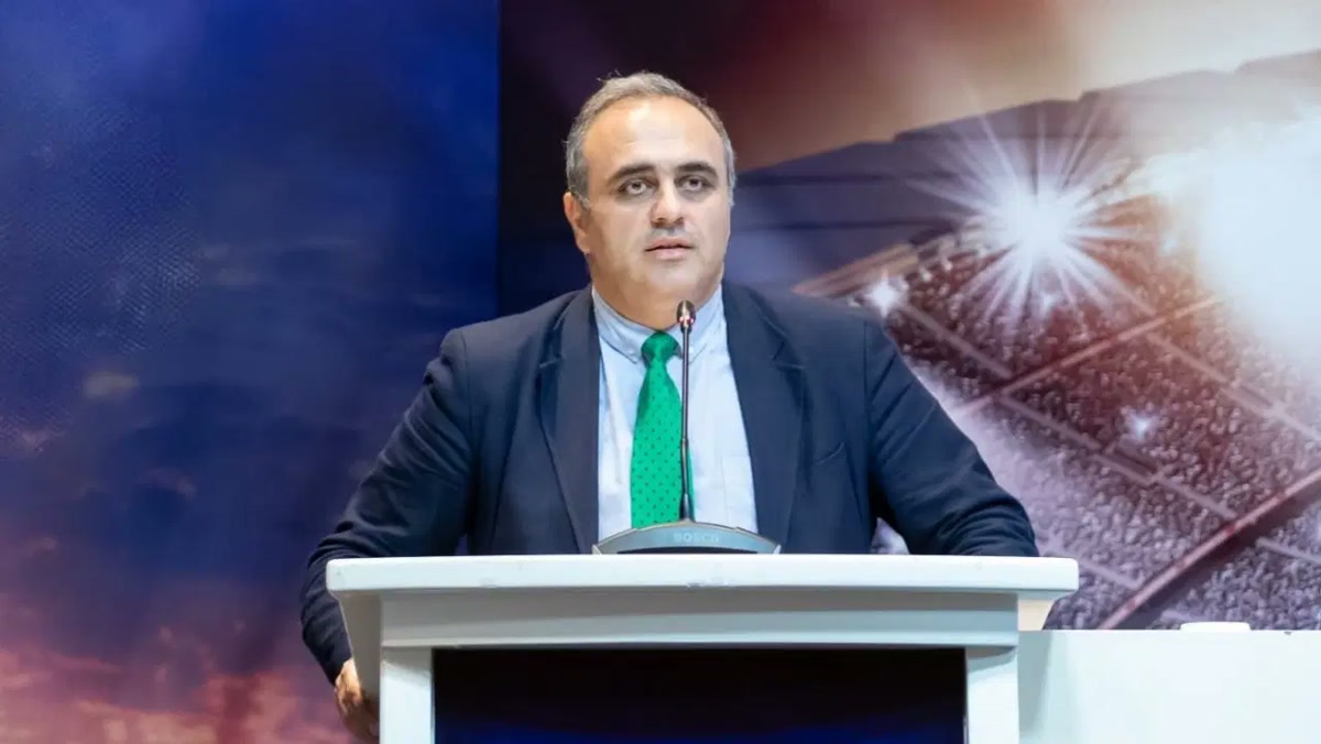 Ural Aküzüm, yeşil kravatla kürsü ve mikrofon başında, sahne fonunun önünde; 1280x720, yazısız temiz kare.