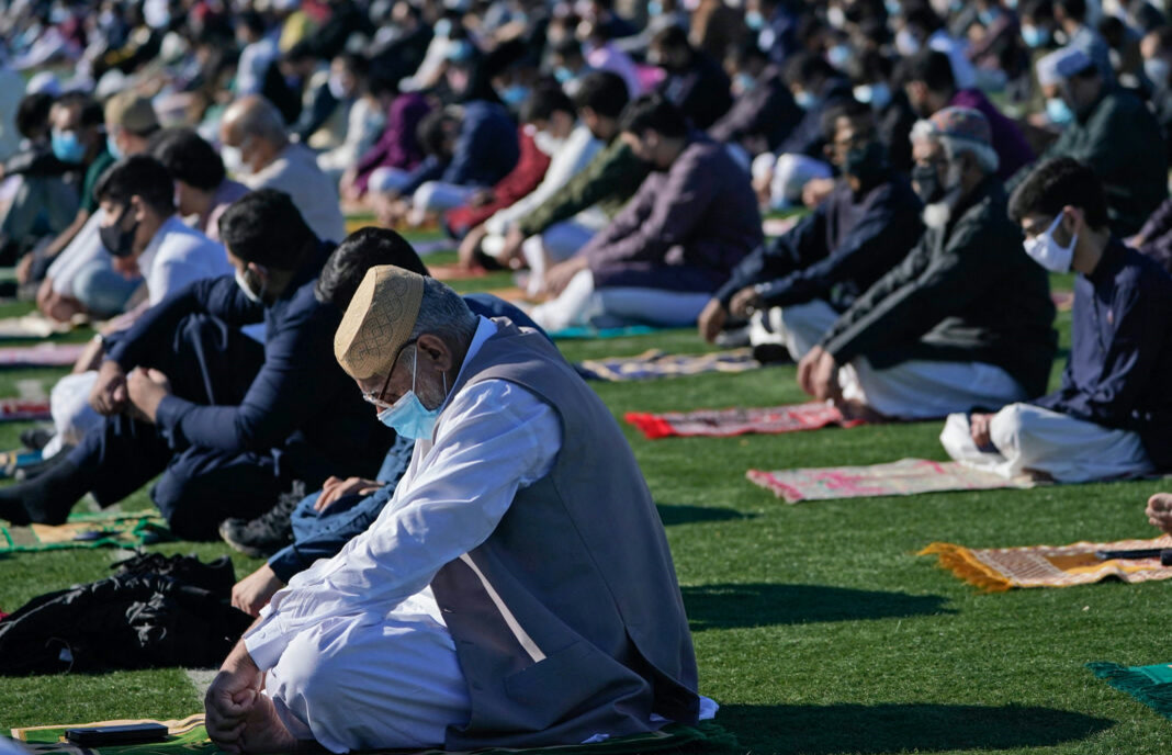 Açık havada çim sahada bayram namazı kılan kalabalık cemaat; ön planda takkeli, maskeli yaşlı bir adam, arkada sıralı cemaat ve seccadeler. 1068x687, net, yazısız. Gövde için bayram görseli (fotoğraf sahibi ve çekim yeri doğrulanmadı).