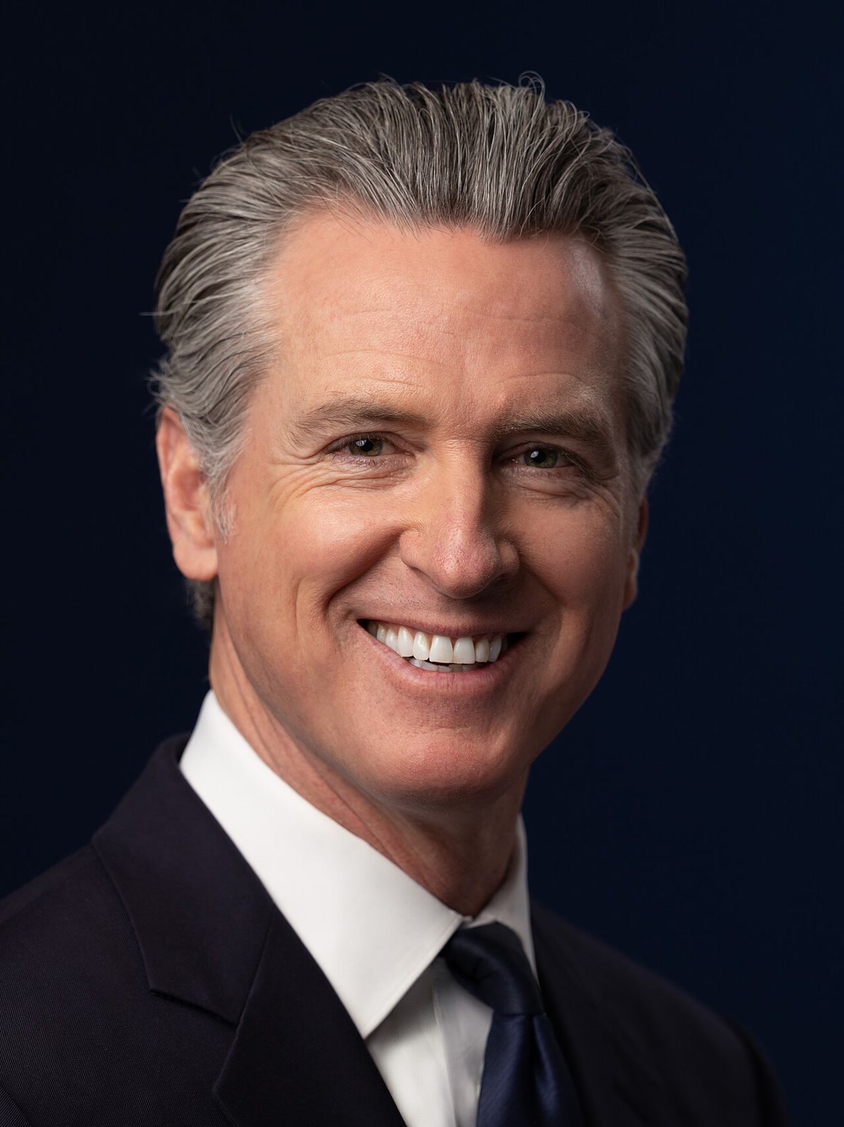 Kaliforniya Valisi Gavin Newsom'un 2026 resmî portresi, koyu lacivert fonda gülümseyen yakın plan; dikey (1280x1708), temiz, yazısız. Vali için gövde görseli.