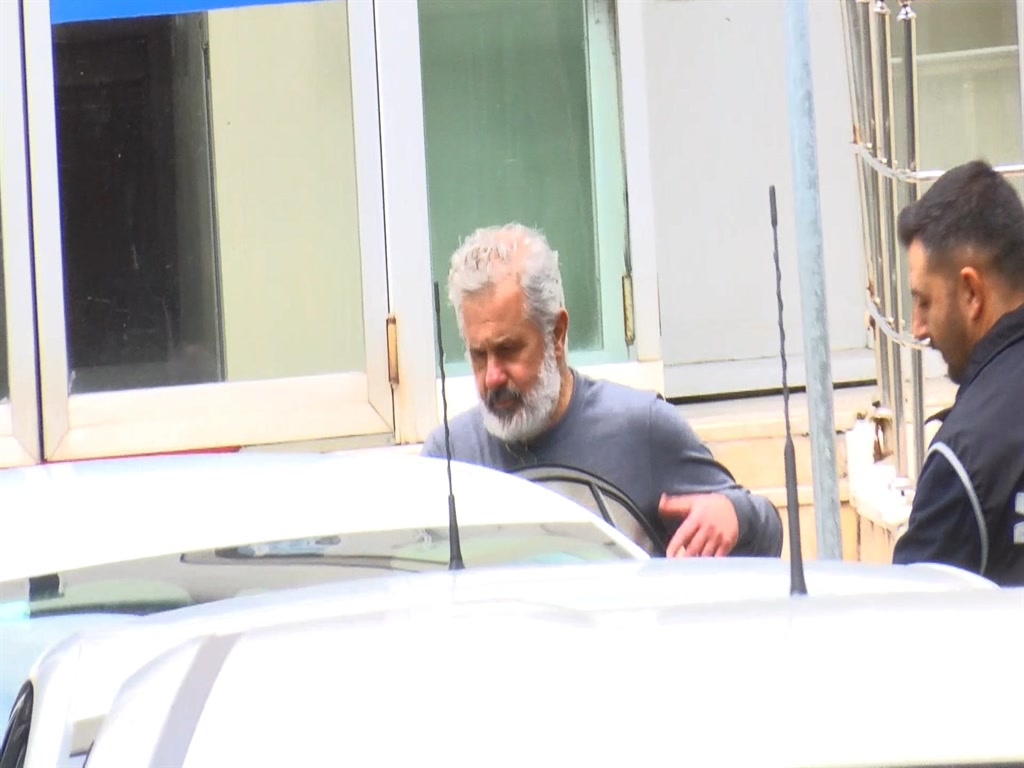 Gri sakallı bir şüphelinin polis eşliğinde yürütüldüğü, binanın önünden çekilmiş saha karesi (1024x768). Yazısız, kolajsız gerçek görüntü; haberin kendi görseli ancak çözünürlüğü orta, gövde fotoğrafı olarak uygun.
