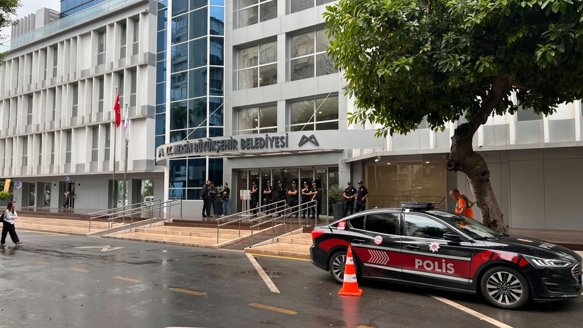 1600x900 temiz fotoğraf: Mersin Büyükşehir Belediyesi binasının cephesi, önünde polis aracı ve giriş kapısında polis ekipleri; tabela tam görünüyor.